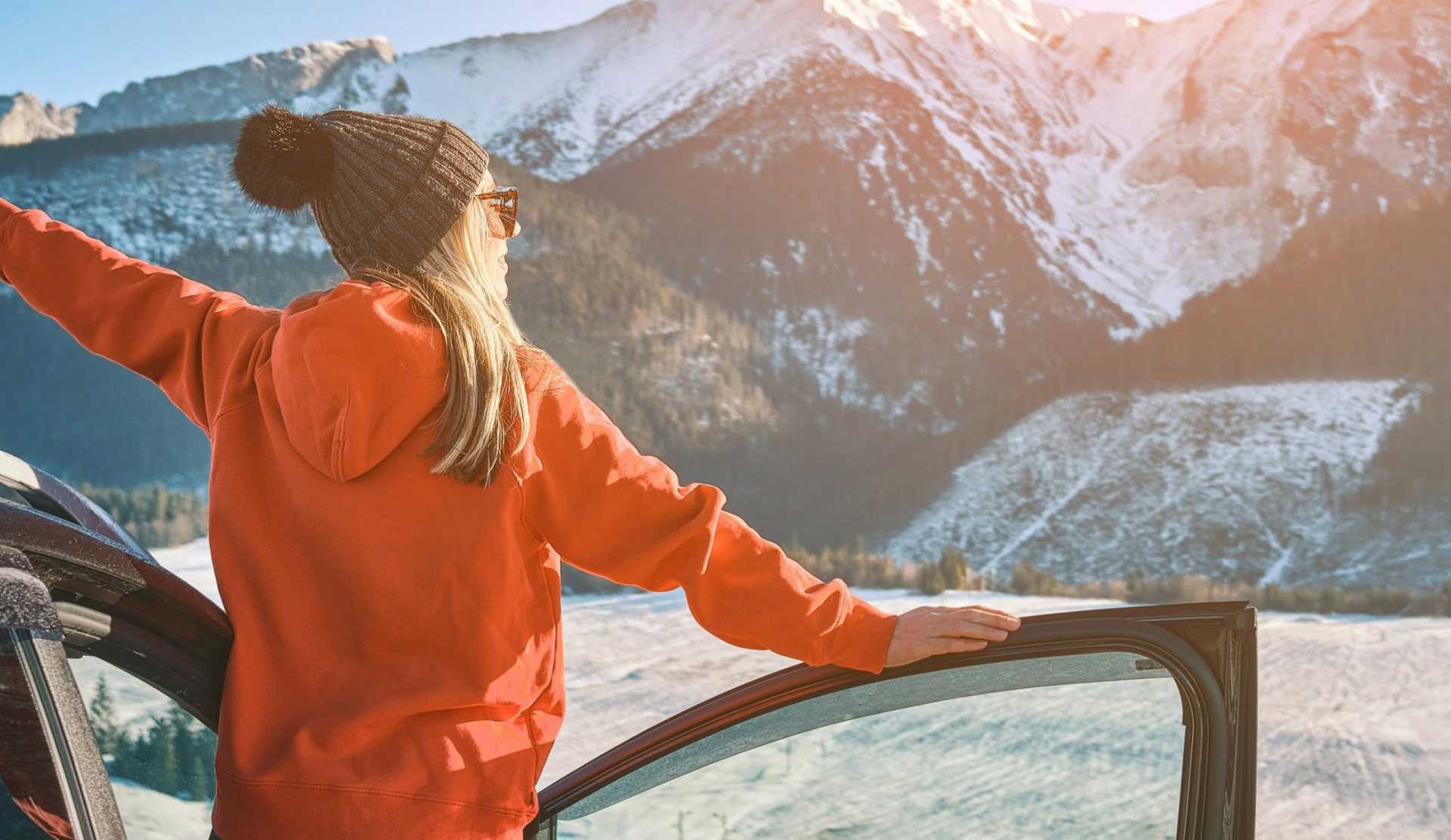 Woman in snow gear leaning out of open car door in awe of snowcapped mountains