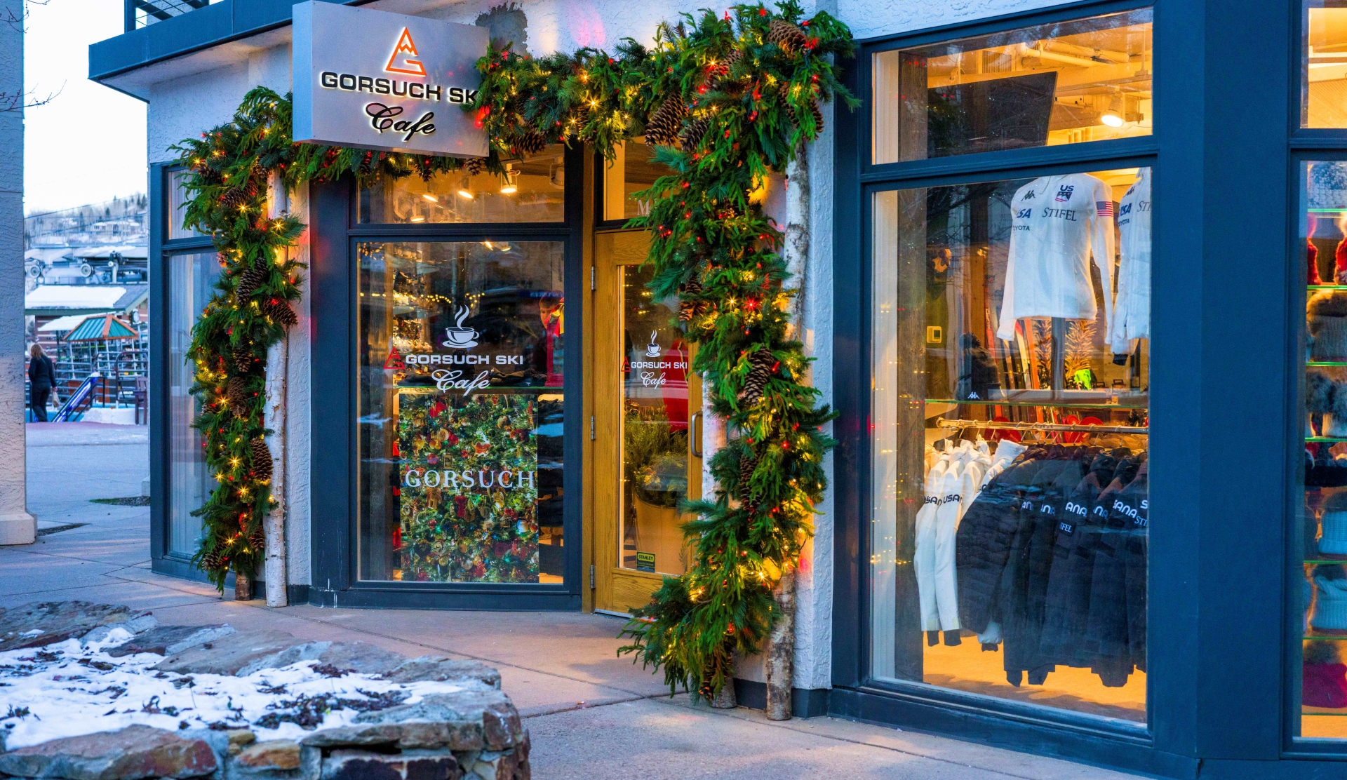 A storefront of Gorsuch Ski Cafe decorated with festive garlands and lights, showcasing winter apparel through large windows.