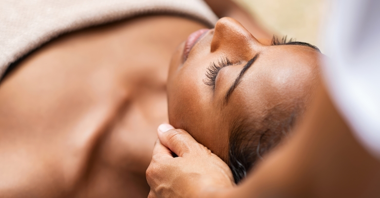 Beautiful African woman getting face massage in beauty spa. Black girl with closed eyes relaxing in outdoor spa while getting head massage. Serene woman relaxing outdoor in a beauty center.